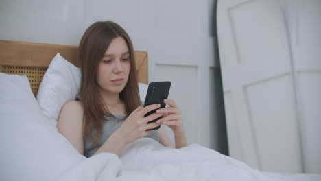 Morgens-Sitzt-Eine-Frau-Im-Schlafzimmer-Und-Lehnt-Sich-An-Die-Weiche-Lederlehne-Des-Bettes.-Schreiben-Sie-Nachrichten-Mit-Ihrem-Smartphone,-Nutzen-Sie-Apps,-Um-Informationen-Zu-Erhalten
